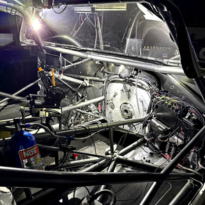 A stripped-out race car interior with a roll cage, exposed engine components, wiring harnesses, and an NOS blue gas bottle in a workshop setting.