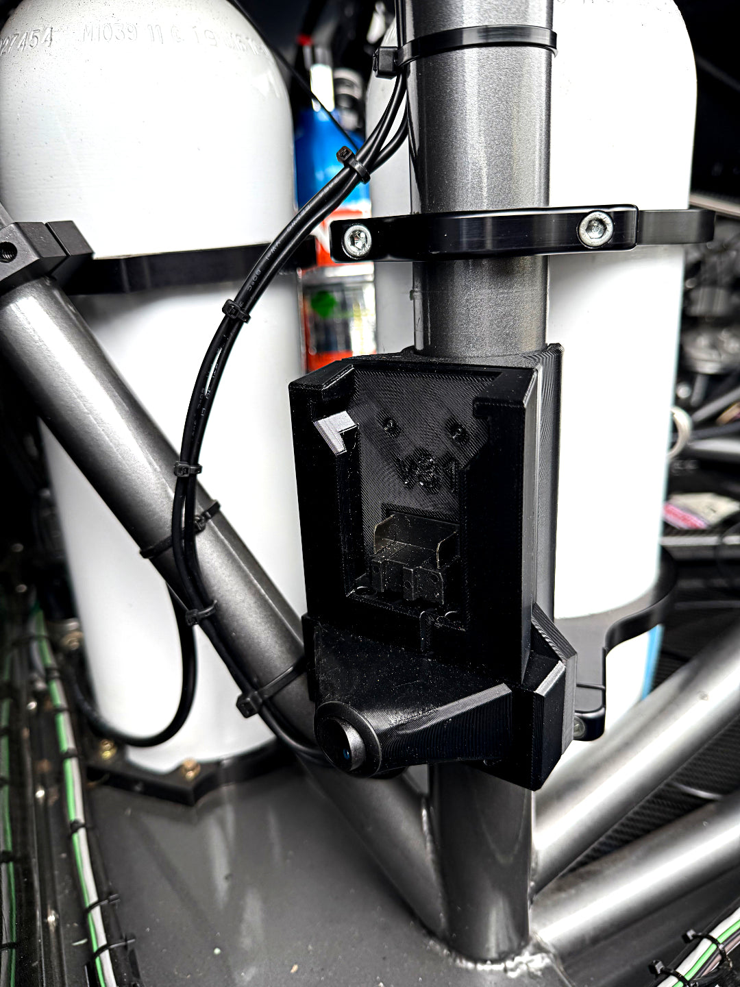 A close-up of a black plastic mounting bracket attached to a silver metal frame, with white cylindrical tanks and black wiring visible in the background.