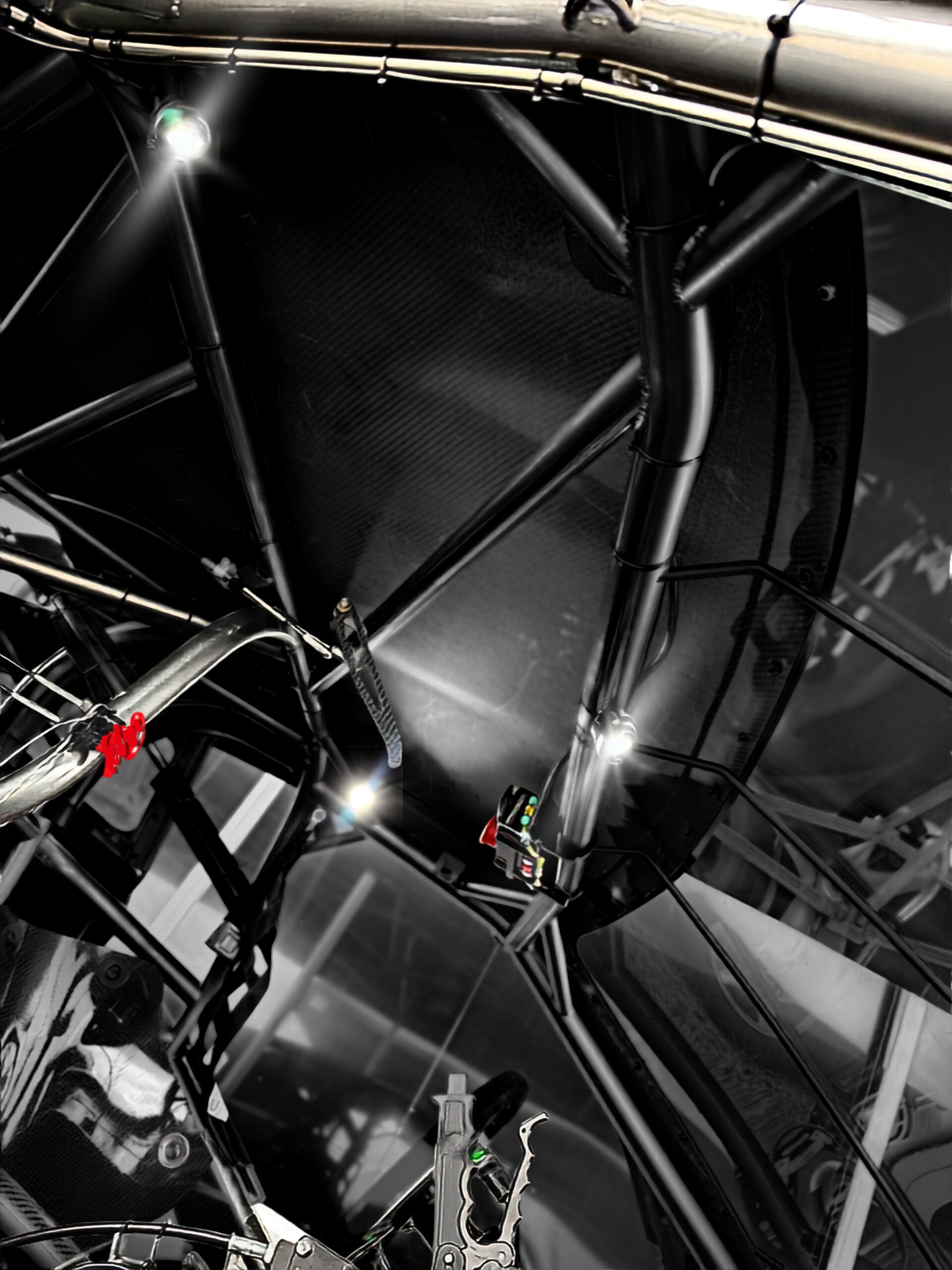 Close-up of a black carbon-fiber roll cage inside a race car cockpit, with bright overhead lights reflecting off glossy metal and frame tubes.