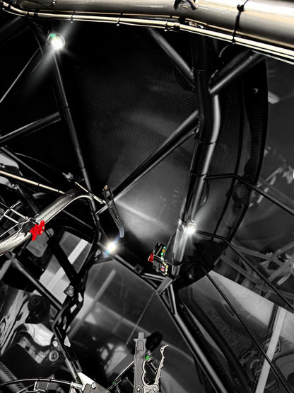 Close-up of a black carbon-fiber roll cage inside a race car cockpit, with bright overhead lights reflecting off glossy metal and frame tubes.