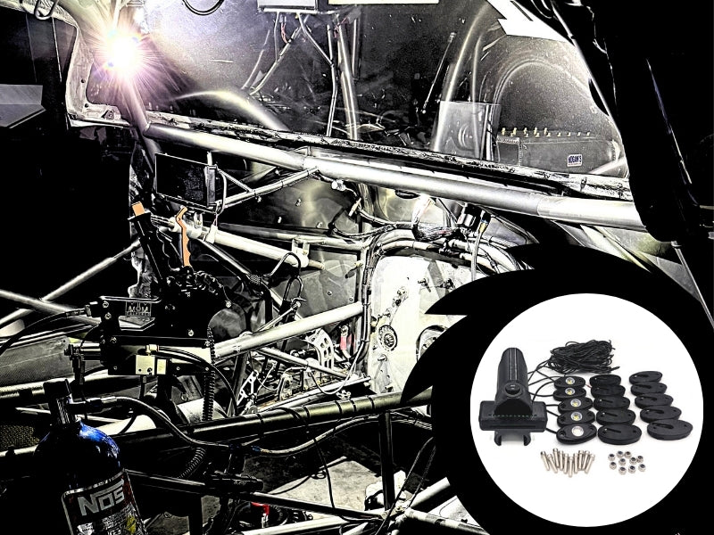 A black automotive sound deadening kit shown in a circular inset overlay, featuring a foam panel, adhesive strips, mounting hardware, and a speaker cover, set against a background of a stripped-out race car frame with a bright work light on.